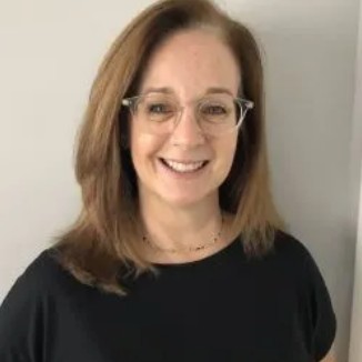 Smiling woman with shoulder-length light brown hair and glasses, wearing a black top indoors against a light wall.