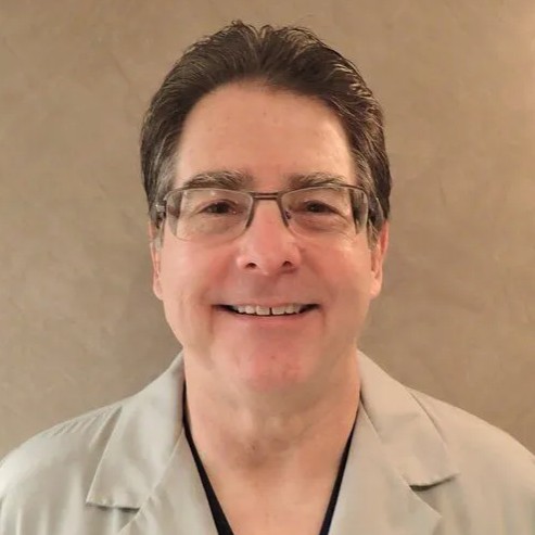 Head-and-shoulders portrait of a smiling person with short dark hair and glasses wearing a light gray blazer in front of a neutral wall.